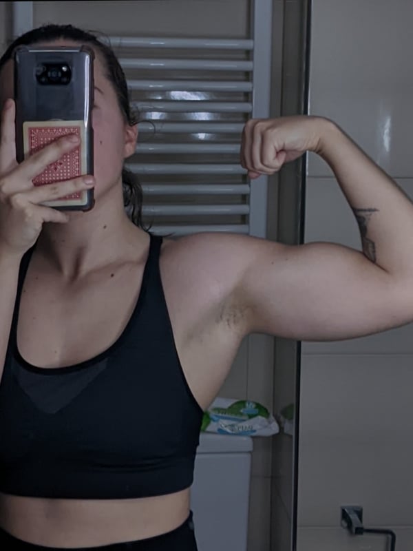Woman flexing her muscular arms in a black sports bra, holding her phone for a mirror selfie in a bathroom setting.