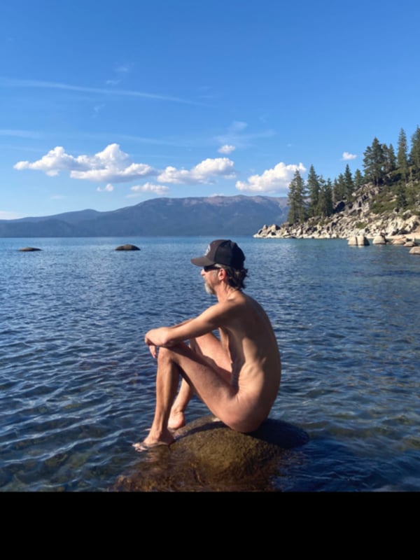 Person sitting nude on a rock by a lake with mountains, trees, and clear blue sky in the background.