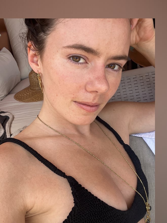A young woman taking a selfie in a bedroom, wearing a black crochet top, with pillows and a hat visible nearby.