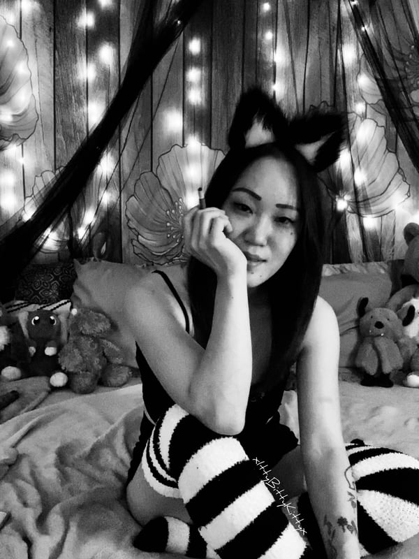 A woman with cat ears and striped stockings sitting playfully on a bed surrounded by fairy lights and stuffed animals in a black and white photo.