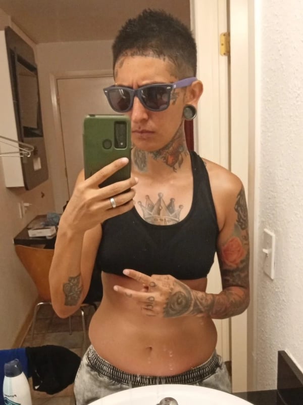 A woman with short hair, sunglasses, and extensive tattoos poses in a sports bra and shorts in a bathroom mirror selfie.