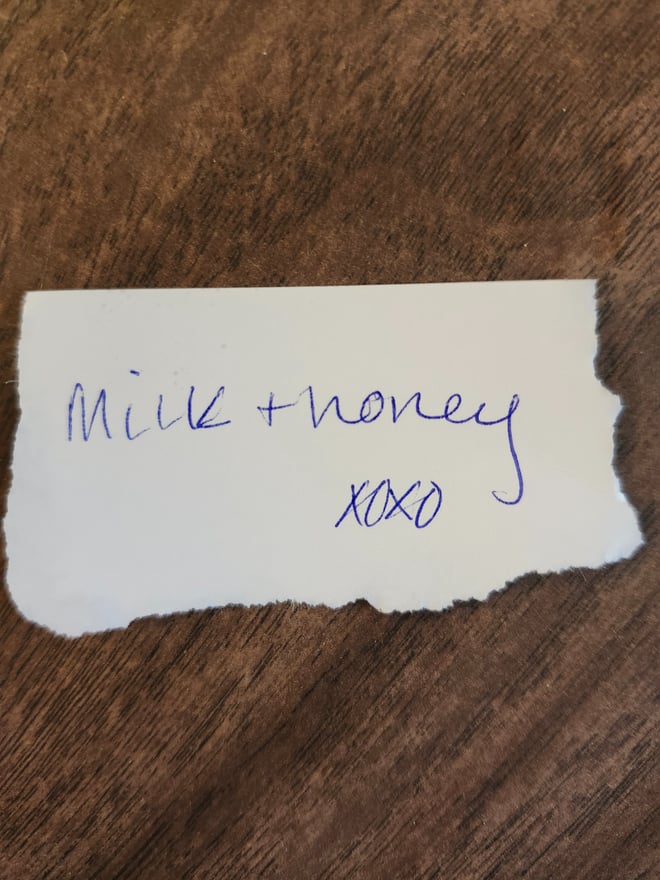 A handwritten note on torn paper reading 'Milk + honey' with 'xoxo' placed on a wooden surface.