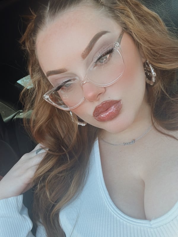 Close-up portrait of a young woman with long wavy brown hair styled in pigtails, wearing clear glasses and hoop earrings, smiling softly in a car.