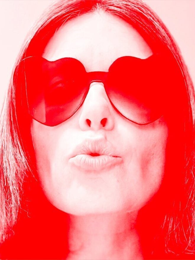 Close-up of a woman with long dark hair wearing red heart-shaped sunglasses, blowing a kiss, in a vibrant red-toned image.