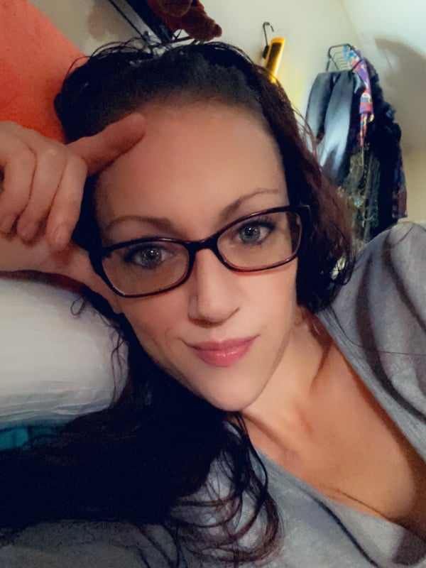 A woman with glasses and long dark hair smiles while reclining on a bed in a cozy room with toys and clothes in the background.