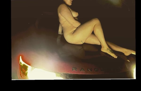 A woman sitting nude on the hood of a luxury car in a dark, dramatic setting with glowing lights.