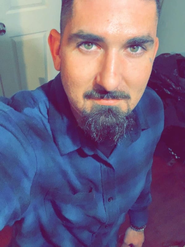A man with dark hair, green eyes, and a beard takes a selfie in a blue plaid shirt, smiling indoors.