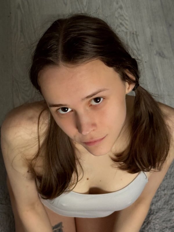 Young woman with brown hair in pigtails looking down at the camera while sitting indoors on a wooden floor.