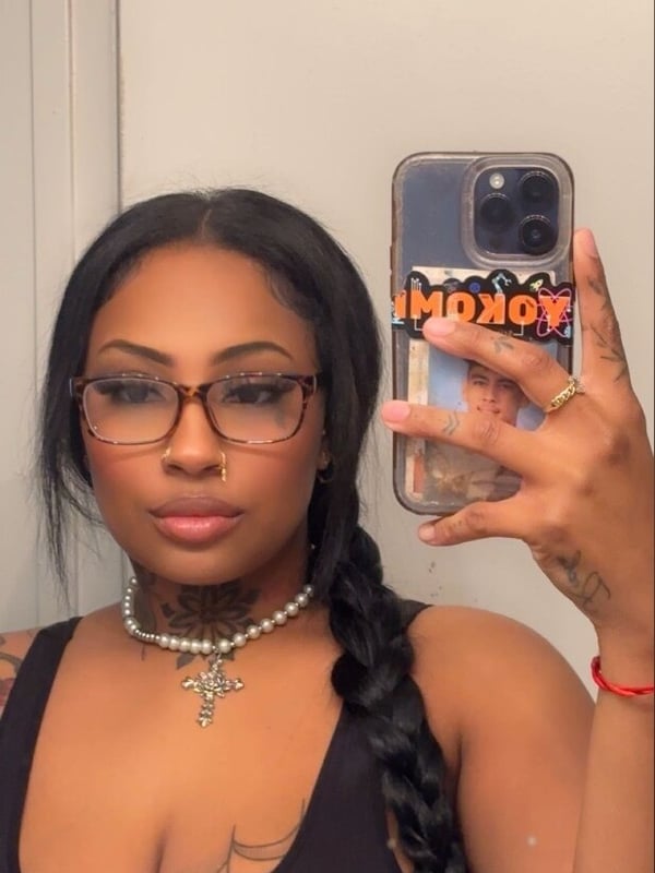 A woman taking a selfie in glasses and a necklace while holding a phone.