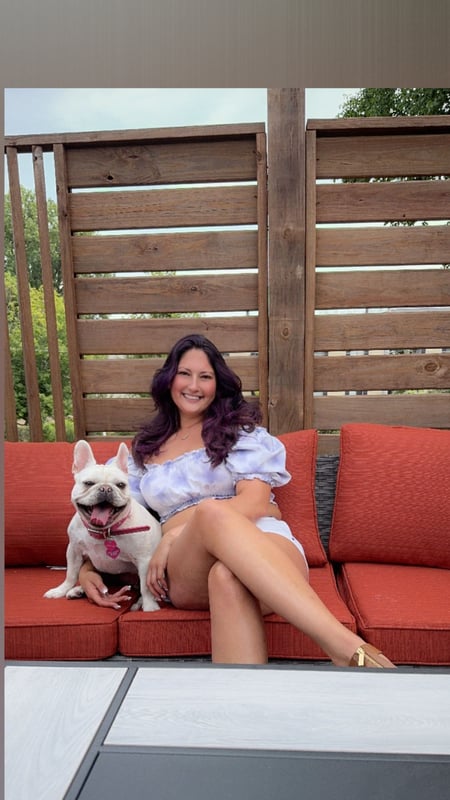 Woman with long wavy hair sitting outdoors on red cushions with a white bulldog, smiling against a wooden fence background.