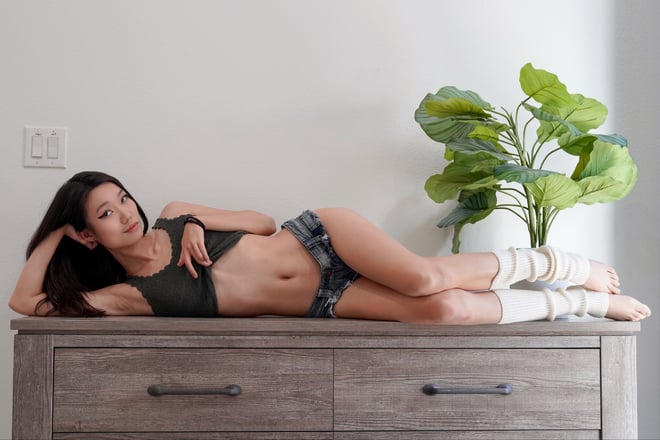 A woman with long dark hair reclining in casual attire on a wooden cabinet beside a leafy houseplant.