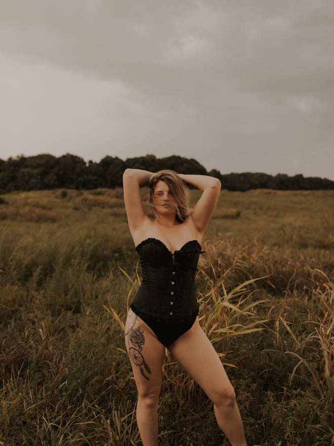 Woman in a black corset-style swimsuit posing with arms raised in a field under a cloudy sky.