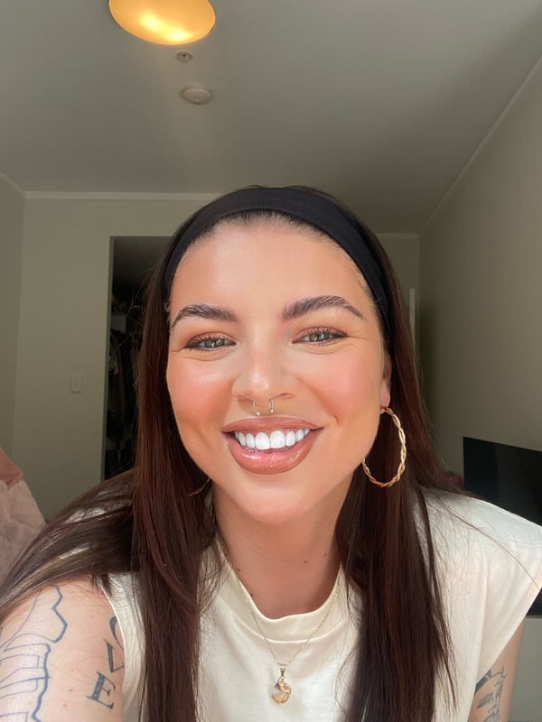 Close-up of a young woman with long dark hair, hoop earrings, and a friendly smile, wearing a white top in a well-lit indoor setting.