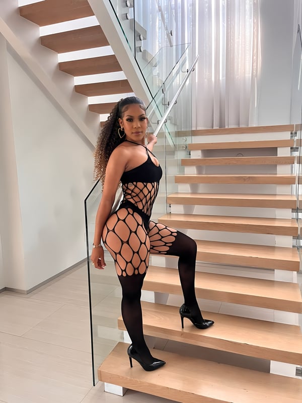 Person posing on wooden stairs in a modern indoor setting with glass railing, wearing a black outfit and high heels.
