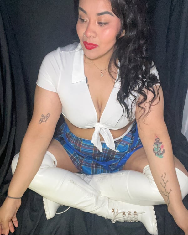 A woman posing seated in a schoolgirl-inspired outfit with a white tied blouse, plaid skirt, and white boots against a dark background.