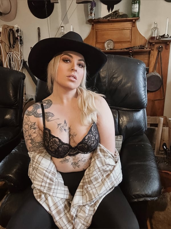 A blonde woman wearing a black cowboy hat, lace top, and plaid shirt, sitting on a leather chair in a vintage room with wooden decor.