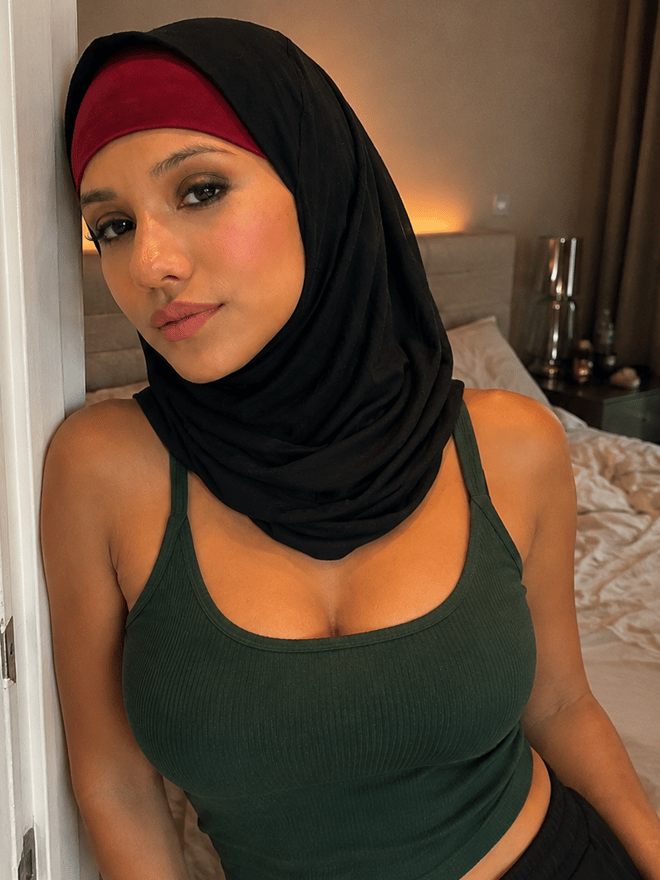 A close-up selfie of a woman wearing a hijab, posing indoors with her hand near her face.