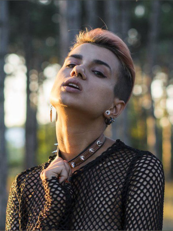 A person with short pinkish hair posing outdoors in a forested area, wearing a mesh top and choker necklace.