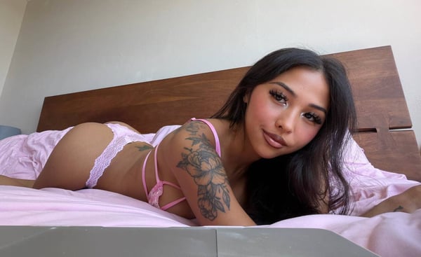 A woman with long dark hair posing on a bed in pink lingerie against a wooden headboard.