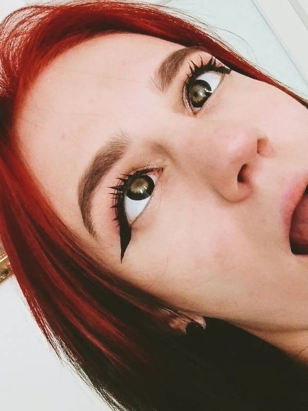 Close-up of young woman with vibrant red hair and green eyes, making a playful expression with tongue out in front of a mirror.