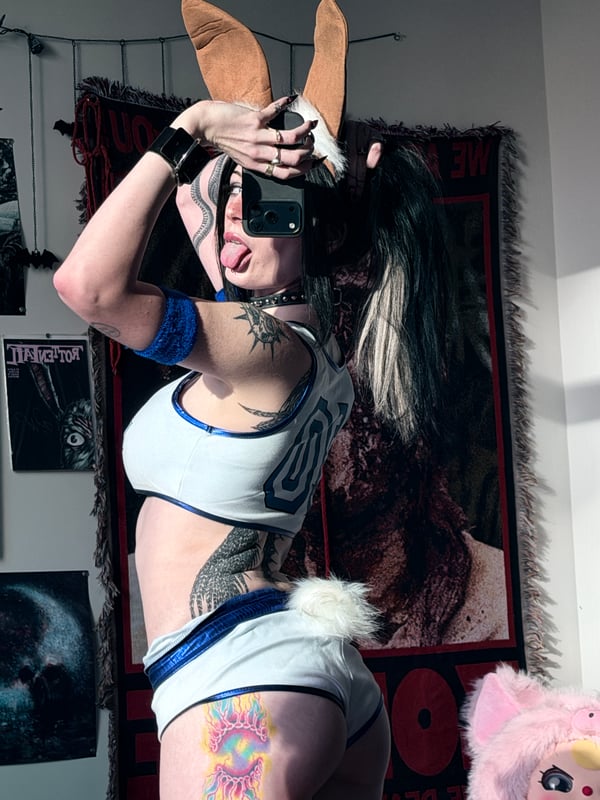 A person with long dark hair and glasses taking a mirror selfie in a cozy room with anime posters and decorations on the wall.