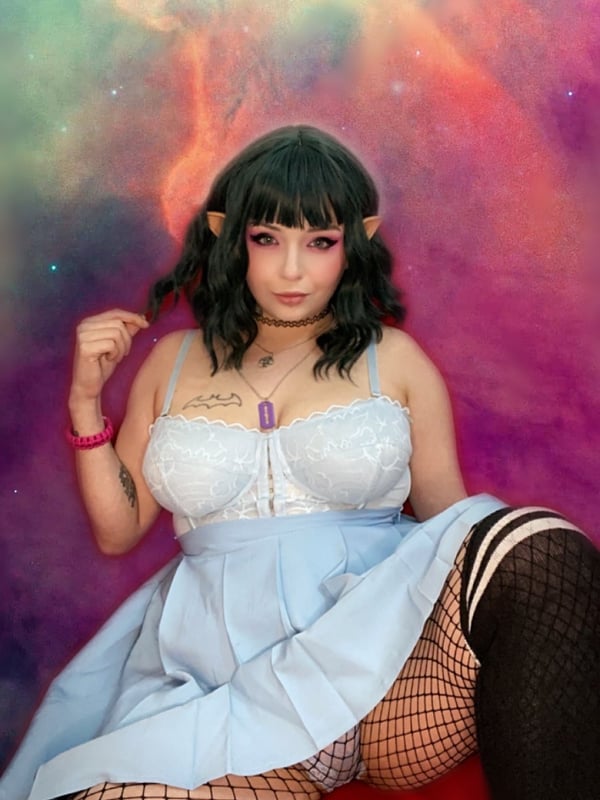 A person with dark hair and elf ears wearing a blue dress and striped stockings, sitting on a red chair against a colorful cosmic background.