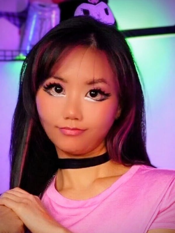 A young woman with striking makeup and dyed hair wearing a pink top and choker, posing confidently against a vibrant purple background with neon lights.
