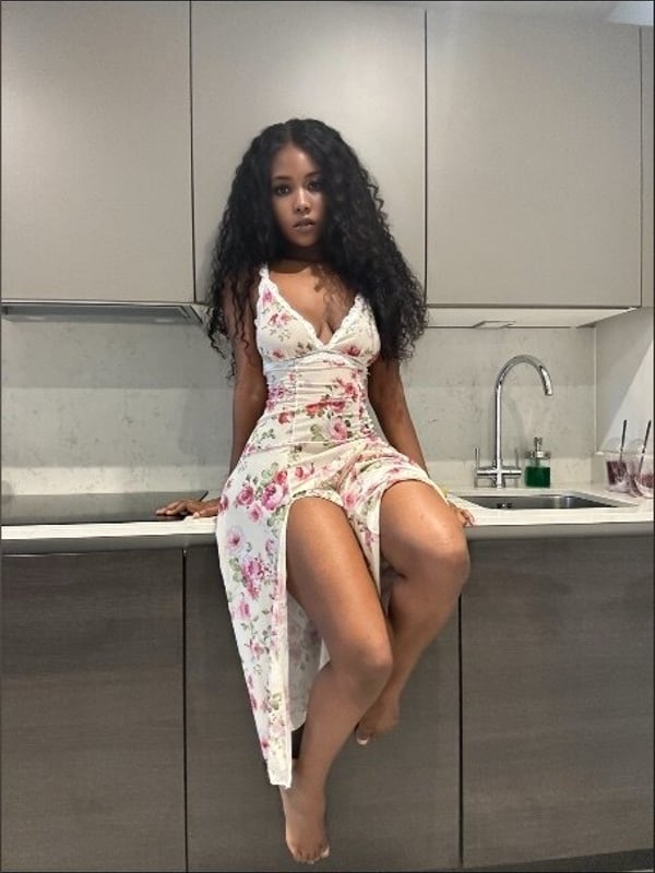 A woman with long curly dark hair wearing a floral dress sitting on a kitchen counter.