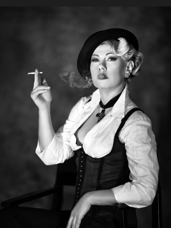 A woman in a stylish black and white outfit with a beret and corset holds a cigarette, seated against a textured backdrop in dramatic lighting.
