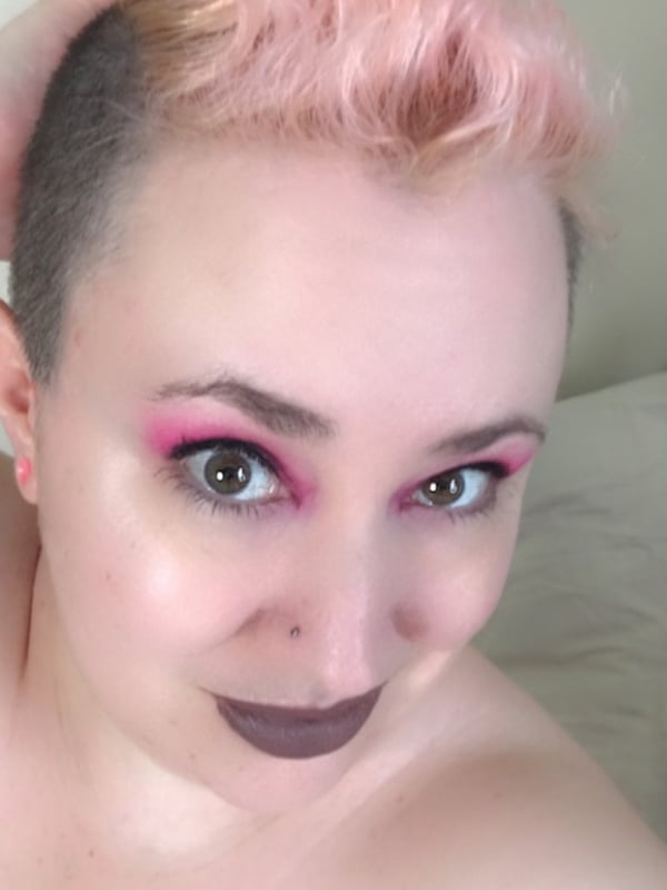 Close-up portrait of a woman with short pink hair, bold makeup, and piercings, smiling at the camera.