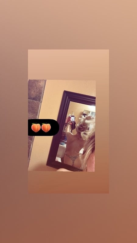 A young woman with long blonde hair taking a mirror selfie in a bedroom, wearing a strapless top and posing playfully.