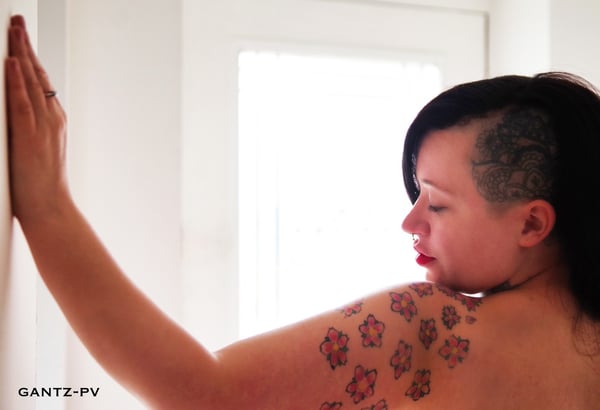 A woman with an undercut hairstyle posing sideways against a white door in a bright room, displaying colorful tattoos on her shoulder.