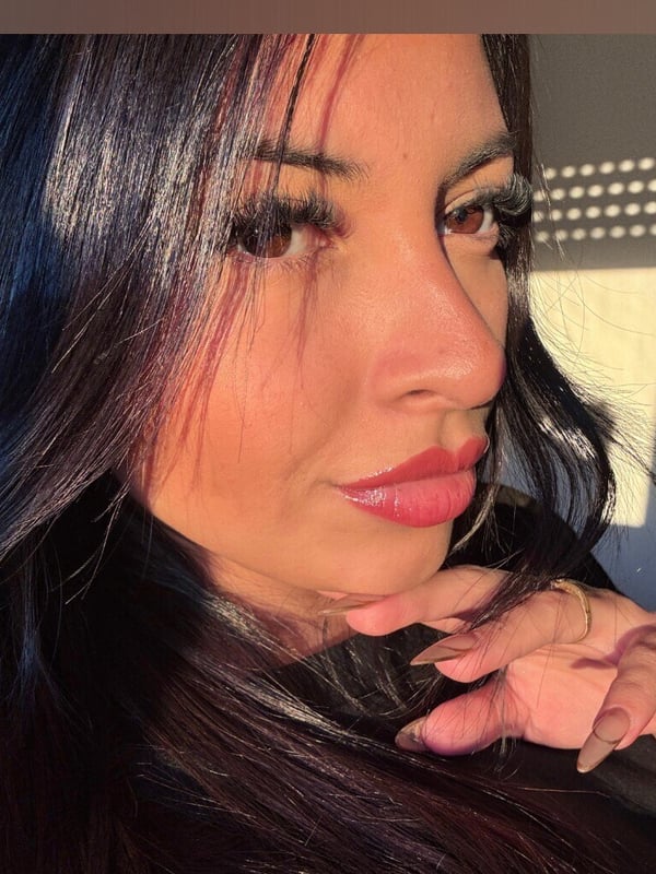 Close-up portrait of a woman with long dark hair, bold makeup, and a subtle smile, illuminated by soft side lighting in an indoor setting.