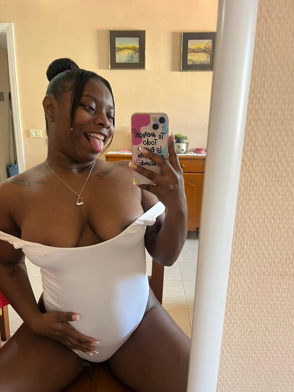 A woman taking a selfie in a white off-shoulder top, sitting with a visible baby bump, smiling in an indoor room.
