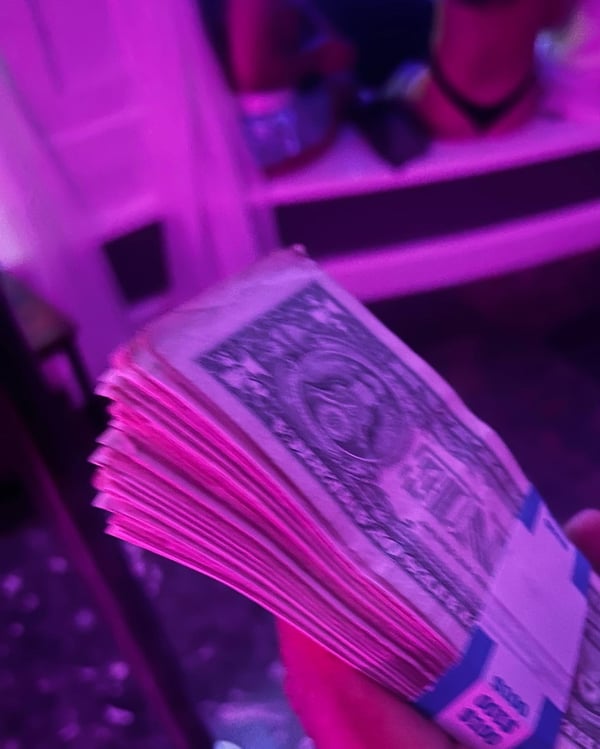 A stack of U.S. dollar bills held in hand, illuminated by vibrant purple lighting in a dimly lit room.