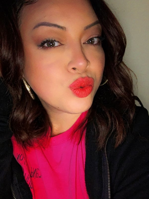 A woman with wavy brown hair and red lipstick making a kiss face, wearing hoop earrings and a pink top under a black jacket.