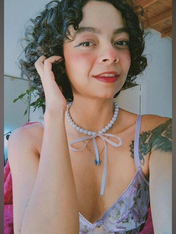 Close-up selfie of a young woman with curly dark hair, smiling with hand near her face, wearing a necklace and floral top in an indoor setting.