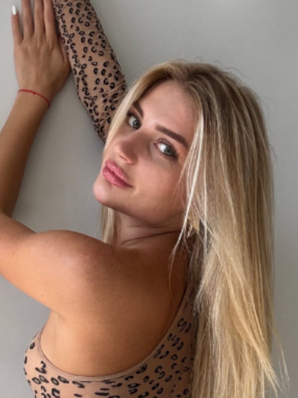 Young woman with long blonde hair wearing a leopard print top, posing with arms raised against a white wall.