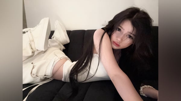 A young woman with long dark hair reclining on a dark couch, wearing a white top and light pants.
