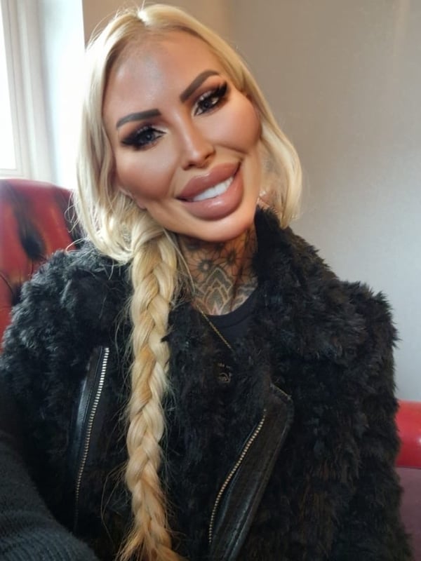 A woman with blonde braided hair and heavy makeup smiles in a stylish black fur jacket and leather outfit inside a warmly lit room.