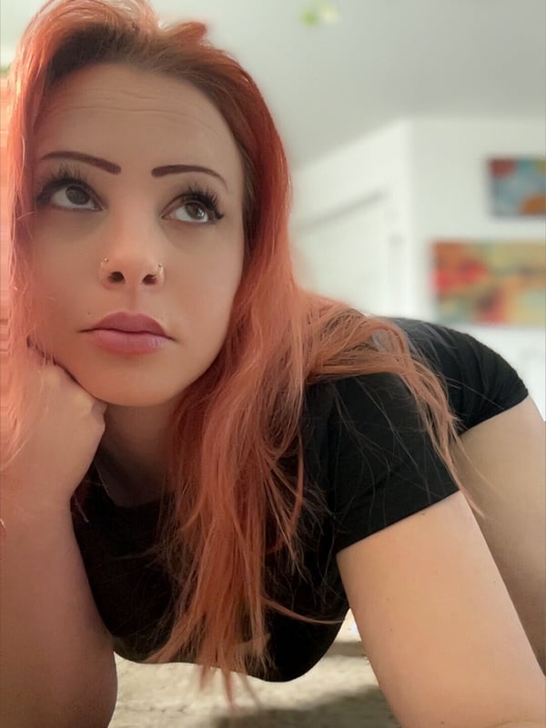 A woman with red hair and bold eyebrows poses on the floor in a black top, resting her chin on her hand in an indoor setting with plants and artwork.