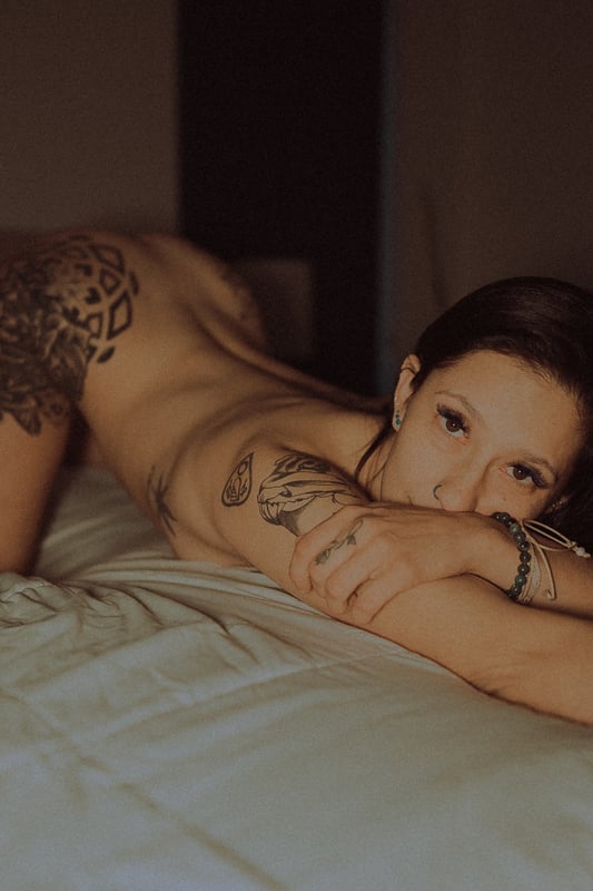 A woman with tattoos reclines on a bed in a dimly lit room, posing confidently with a sultry expression.
