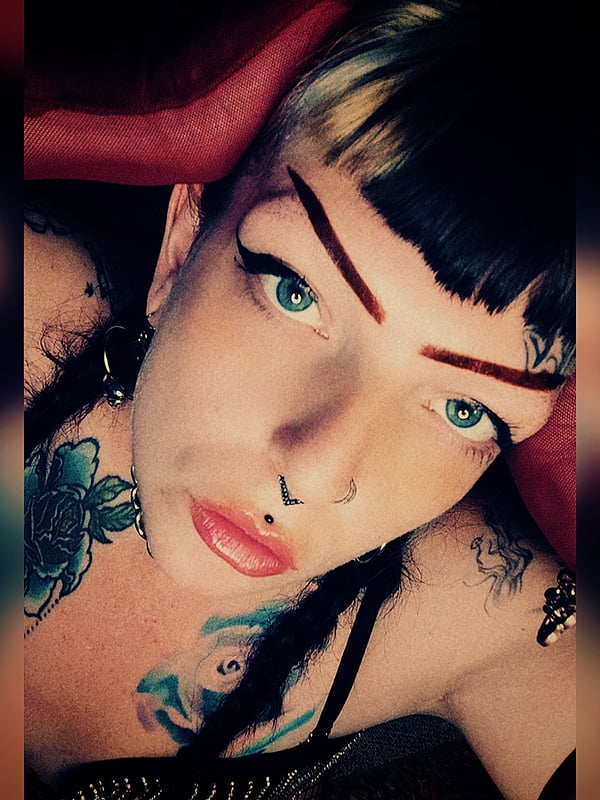 Close-up portrait of a woman with bold makeup, piercings, tattoos, and dyed hair posing confidently.