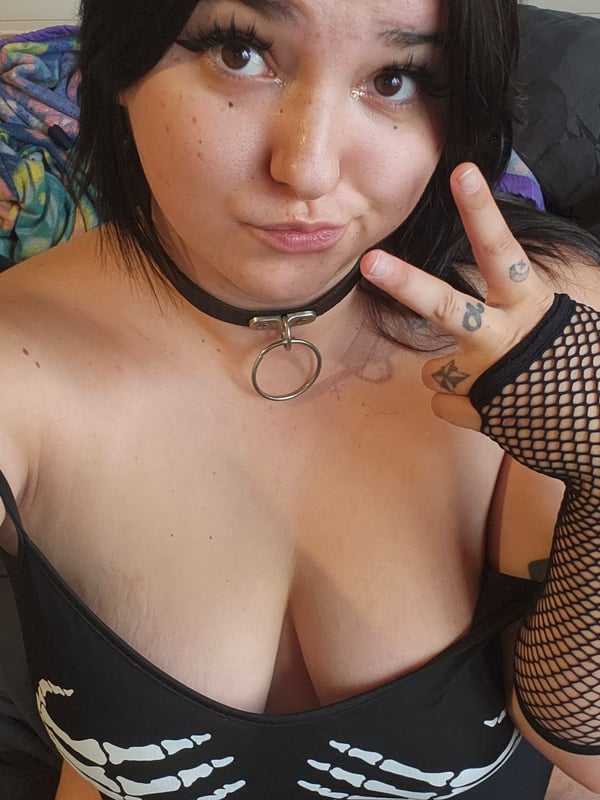A young woman with dark hair and bold makeup makes a playful face with a peace sign, wearing a black choker and mesh arm sleeve in a casual bedroom setting.