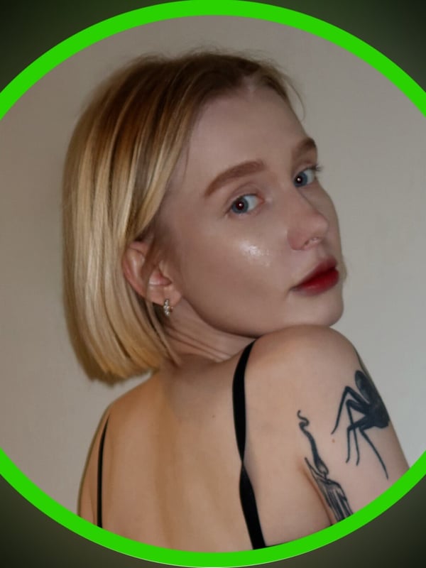 Close-up portrait of a young blonde woman with subtle makeup and tattoos on her shoulder, framed in a green circle against a dark background.