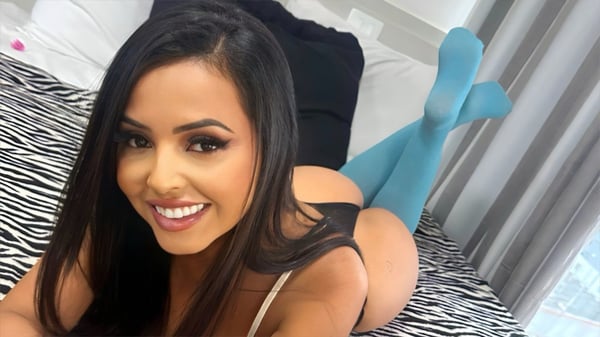 A woman with long dark hair smiling playfully while lying on a bed with zebra print sheets, wearing a black top and blue stockings, in a bedroom with window light.