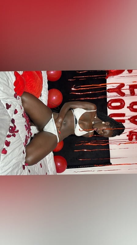 A woman posing on a bed surrounded by red balloons and rose petals in a festive setting.