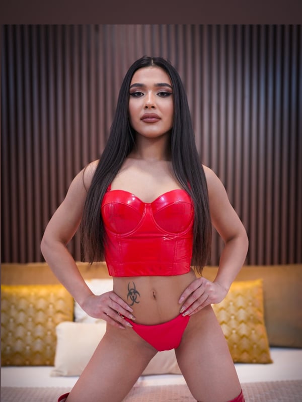 A woman standing with hands on hips in red outfit and high boots against a tufted wall in a bedroom.