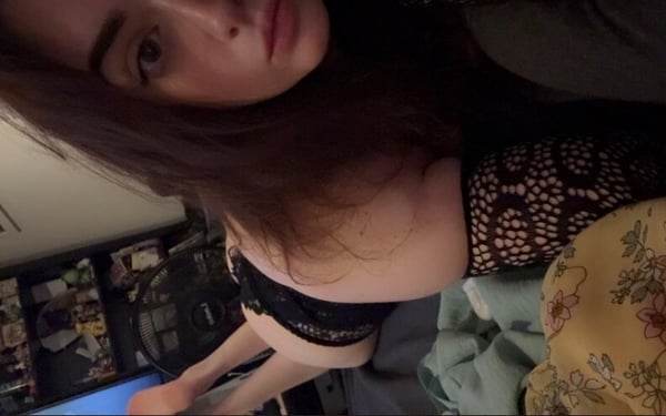Young woman with long brown hair posing in lace topwear in a cozy indoor setting with plants and monitors in the background.