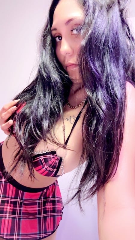 A woman with long dark hair posing confidently indoors, wearing a plaid skirt and top.